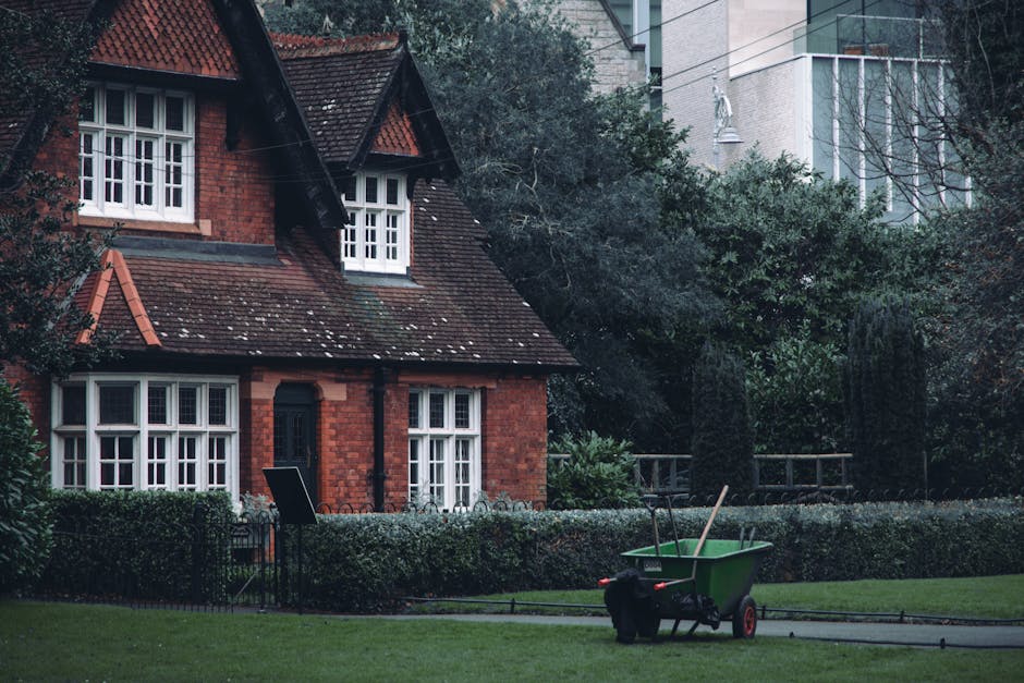 A tidy front garden featuring a well-maintained lawn with neatly trimmed grass, bordered by a low hedge and various shrubs. A wheelbarrow filled with gardening tools, including a rake and a hoe, is po