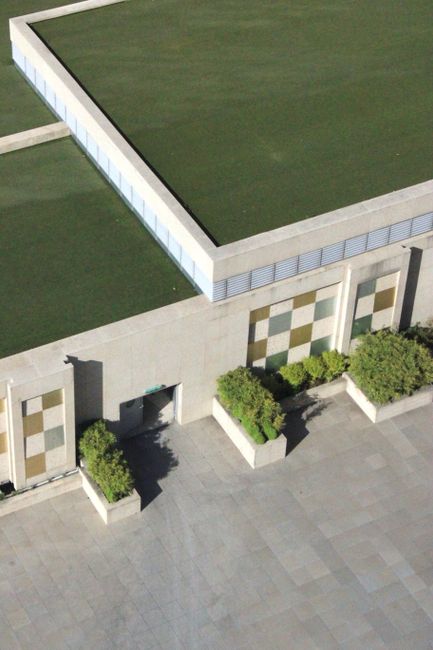 An aerial view of a modern outdoor area featuring a large flat roof covered in well-maintained, lush green grass. The building's exterior wall is decorated with a geometric pattern of beige, brown, an